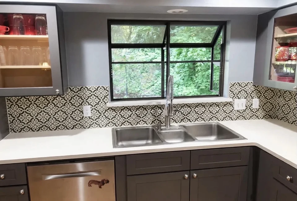 Kitchen sink beneath a dark-framed window, with patterned tile, a pale countertop, dark lower cabinetry, and open shelves.