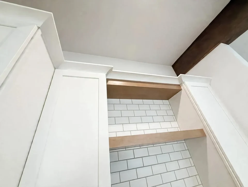 Natural wood open shelves between white cabinets above a white subway-tile backsplash beneath an exposed ceiling beam.