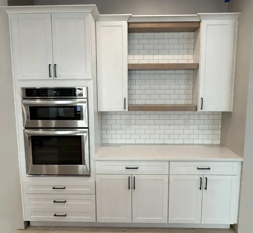 White cabinetry surrounding two stacked stainless steel ovens, with natural wood open shelves and a white tile backsplash.
